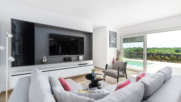 A modern living room with a large flat-screen TV, a grey sofa with pink cushions, a coffee table, and a view of a swimming pool through large sliding doors. - Charming Villa Audrey with an outdoor pool (Umag)