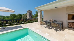 Área na piscina com mesa e cadeiras, um pilar de pedra e vista para uma torre ao fundo.