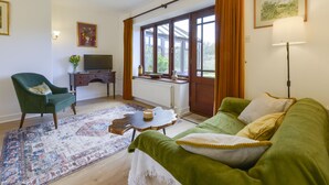 A living room with a green sofa, a green armchair, a wooden coffee table, a patterned rug, a television on a wooden stand, and a window with curtains. - Little Barton (Taunton)
