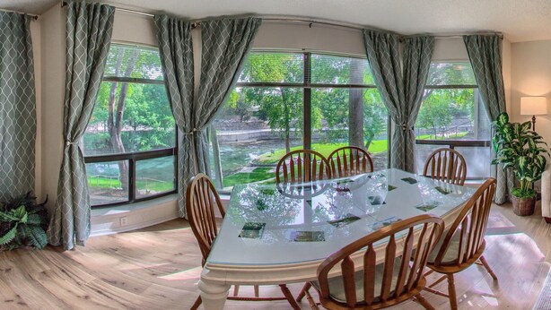 A dining area with a glass table and wooden chairs, large windows with curtains, and a view of greenery outside. - Timeless Tranquility Crc 317 2 Bedroom Condo by RedAwning (New Braunfels)