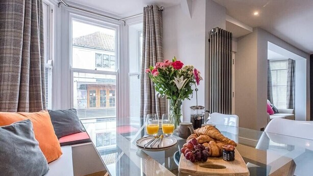 Una sala de estar moderna con una mesa de vidrio, un sofá con cojines, un florero con flores y una bandeja de desayuno con croissants y uvas.