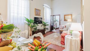 Un salon bien éclairé avec un coin repas, comprenant une télévision, des plantes décoratives et une variété de fruits et de légumes disposés sur la table.