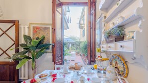 Une table dressée pour un service à thé, avec des pâtisseries et des fruits, une plante en pot, et une vue sur une rue étroite bordée d’immeubles.