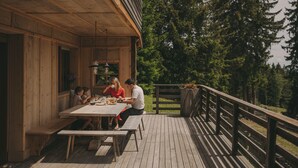 Una terraza de madera con una mesa y bancos, rodeada por una barandilla, con vistas a un área boscosa.