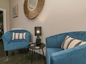 A living room with two blue armchairs, a round mirror, a small table with a lamp, and a radiator. - 7 Belgrave Apartments (Ilfracombe)