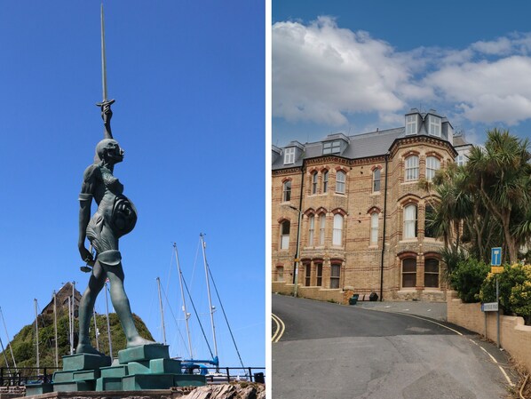 A statue of a figure holding a sword, with boats in the background. The image also shows a building with a curved roof and a street in front. - 7 Belgrave Apartments (Ilfracombe)