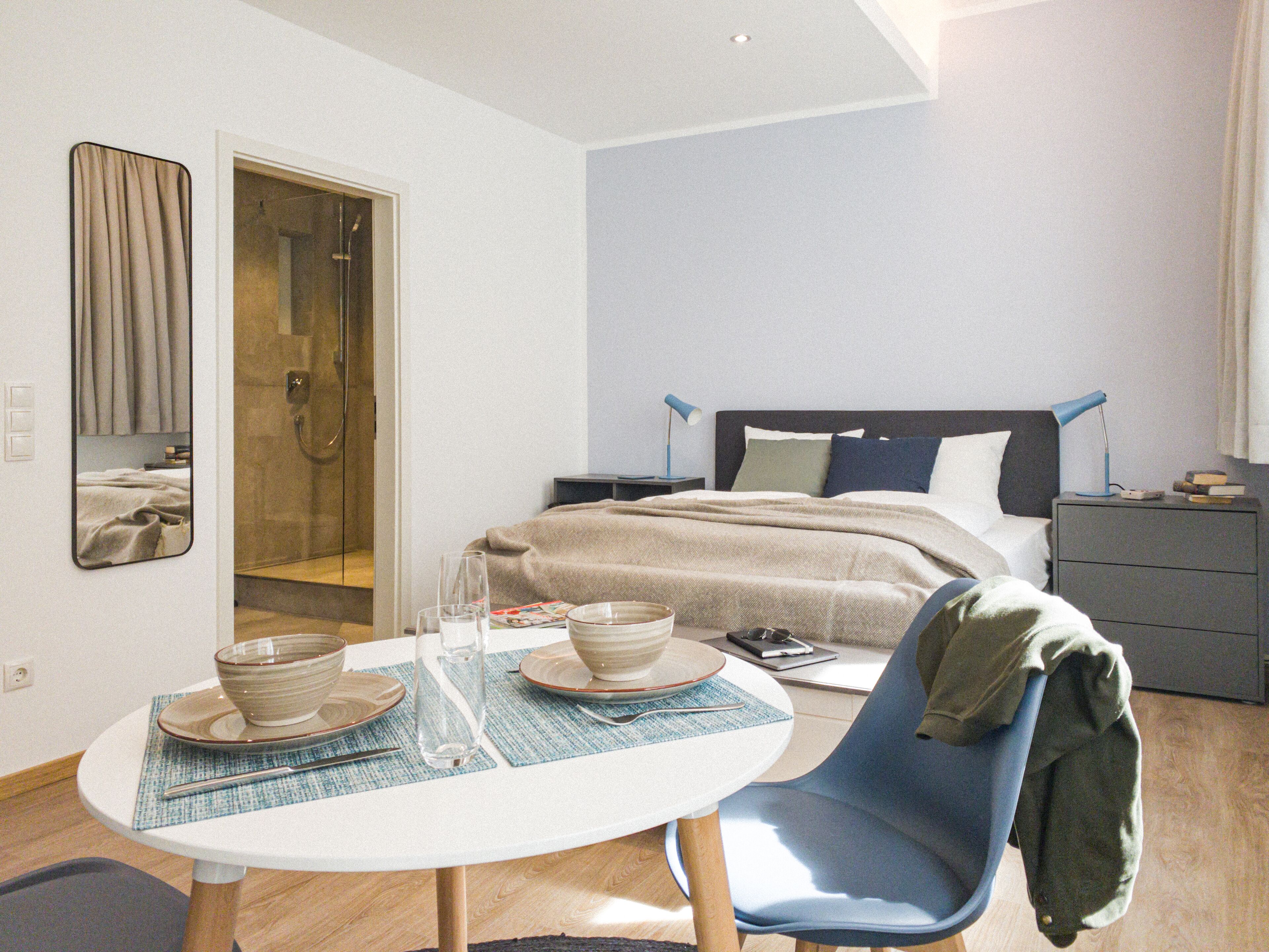 A modern hotel room with a bed, bedside table, and a dining area with a table and chairs.