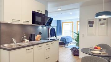 A modern kitchen with white cabinets, a black microwave, and a sink. There is a dining area with a table set for two, a chair, and a plant.