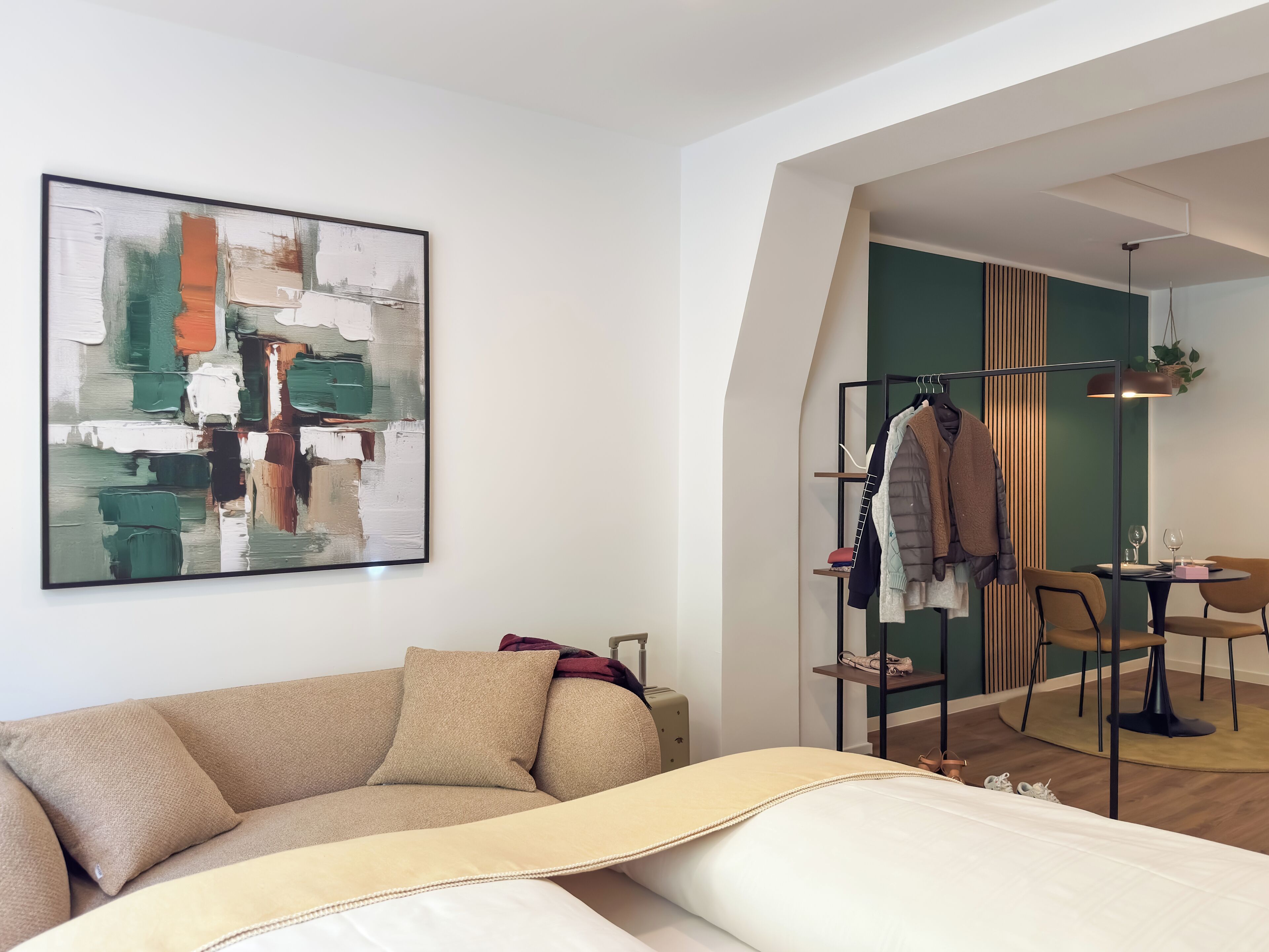 A modern hotel room with a beige sofa, a large abstract painting, a clothing rack, and a dining area with a table and chairs.