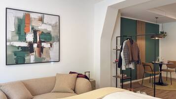 A modern hotel room with a beige sofa, a large abstract painting, a clothing rack, and a dining area with a table and chairs.