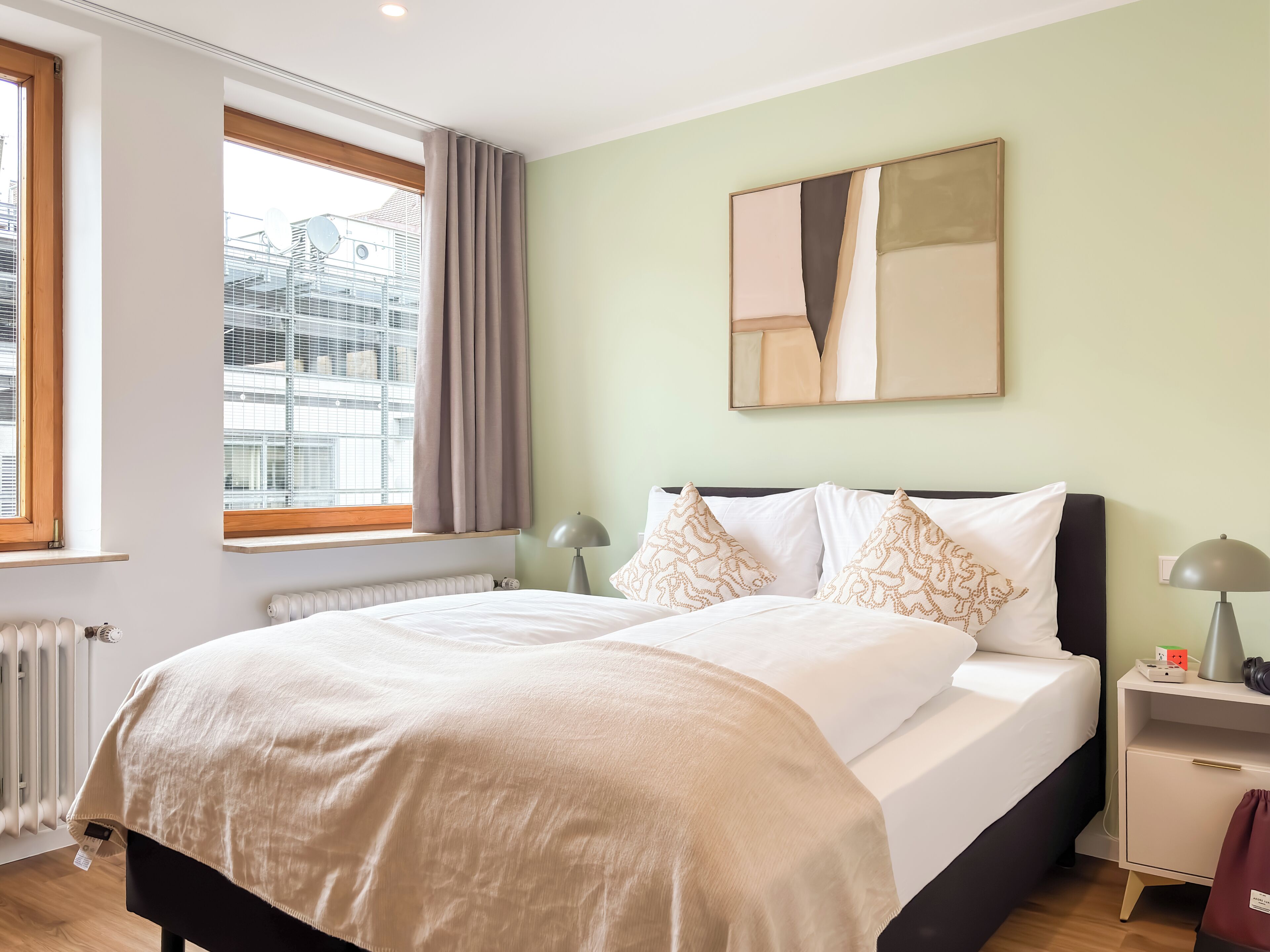 Una cama bien hecha con una manta color beige, una mesita de noche con lámpara y una ventana con cortinas.