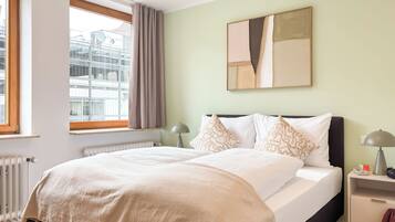 Ein ordentlich bezogenes Bett mit einer beigen Decke, ein Nachttisch mit Lampe und ein Fenster mit Vorhängen.