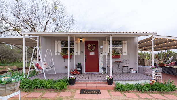 Une petite maison de plain-pied avec une porte rouge, un porche doté d’une balançoire et un paillasson sur lequel est écrit « Bonjour » à l’entrée.