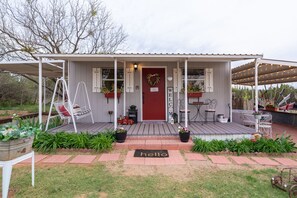 A small, single-story house with a red door, a porch with a swing chair, and a 'hello' mat at the entrance. - Unique Tiny House with Queen Bed, Hot Tub & WIFI in country setting. (Sweetwater)