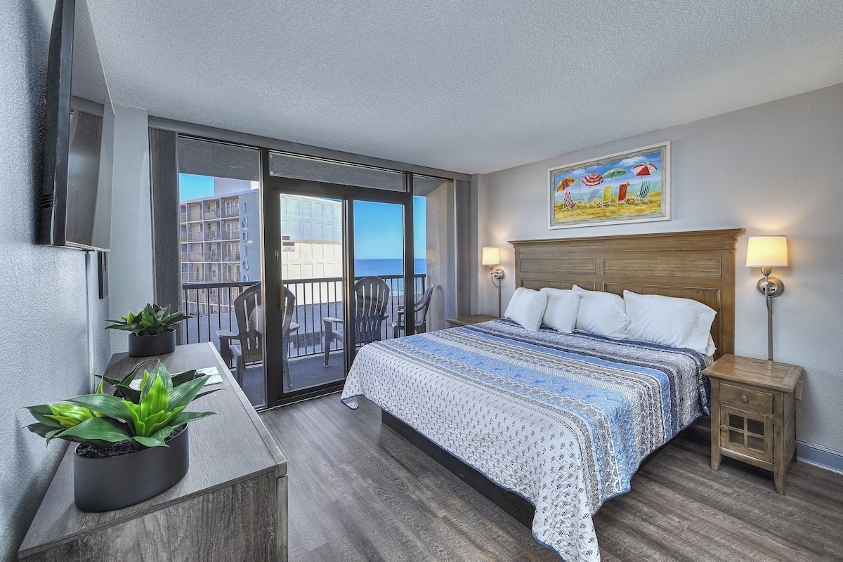 Habitación de hotel con una cama grande, un escritorio con una planta, un balcón con vista al océano y un cuadro en la pared.