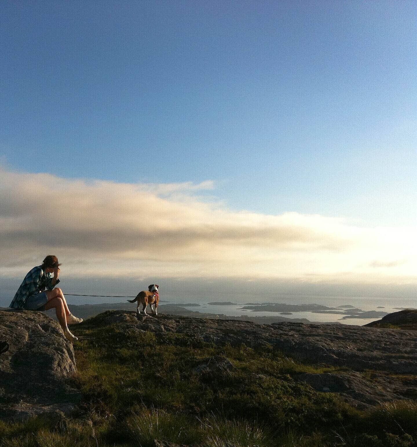 Eine Person sitzt auf einem Felsen mit Blick auf ein Gewässer, ein Hund ist in der Nähe.