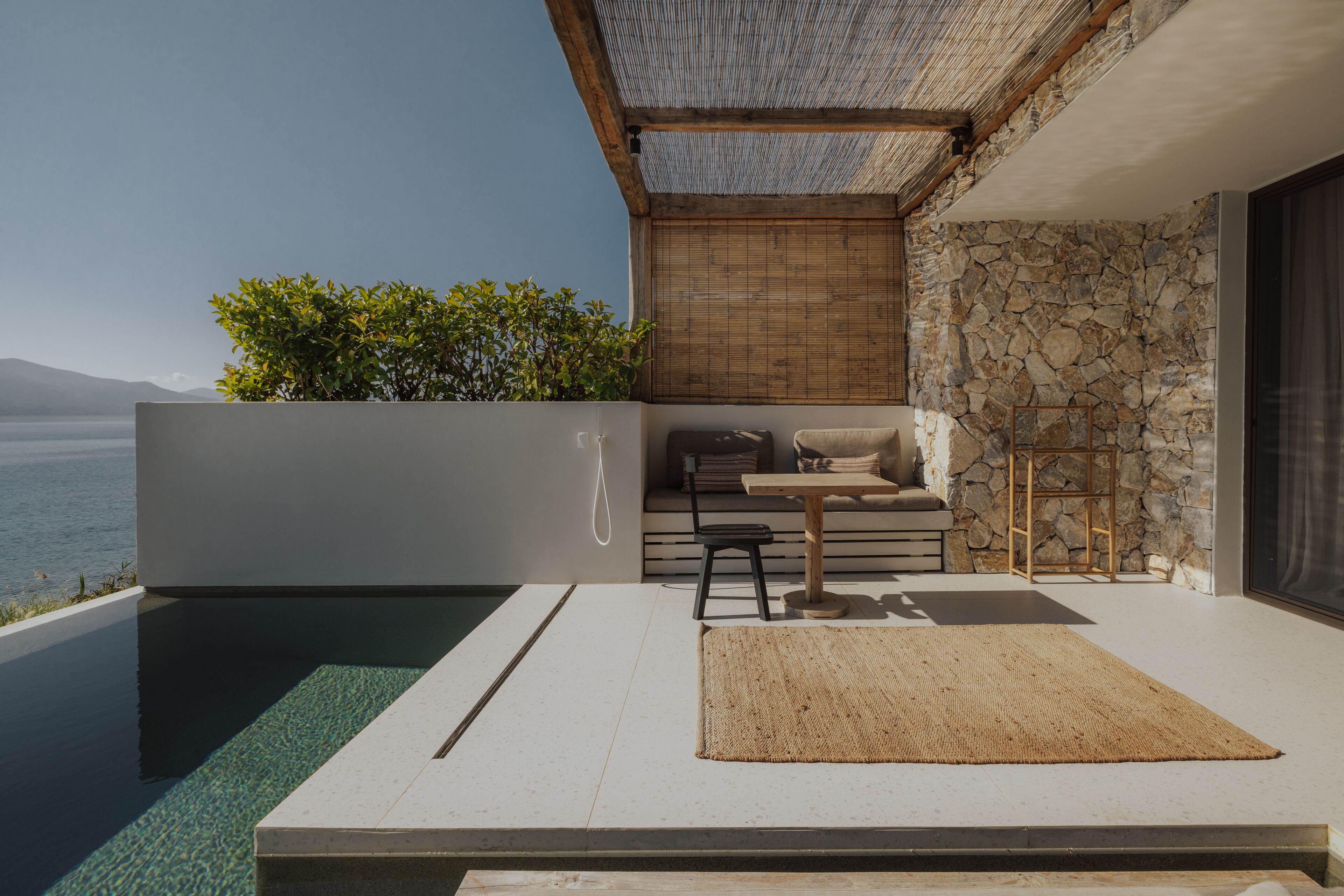 A modern outdoor living space with a pool, a seating area with cushions, and a stone wall.