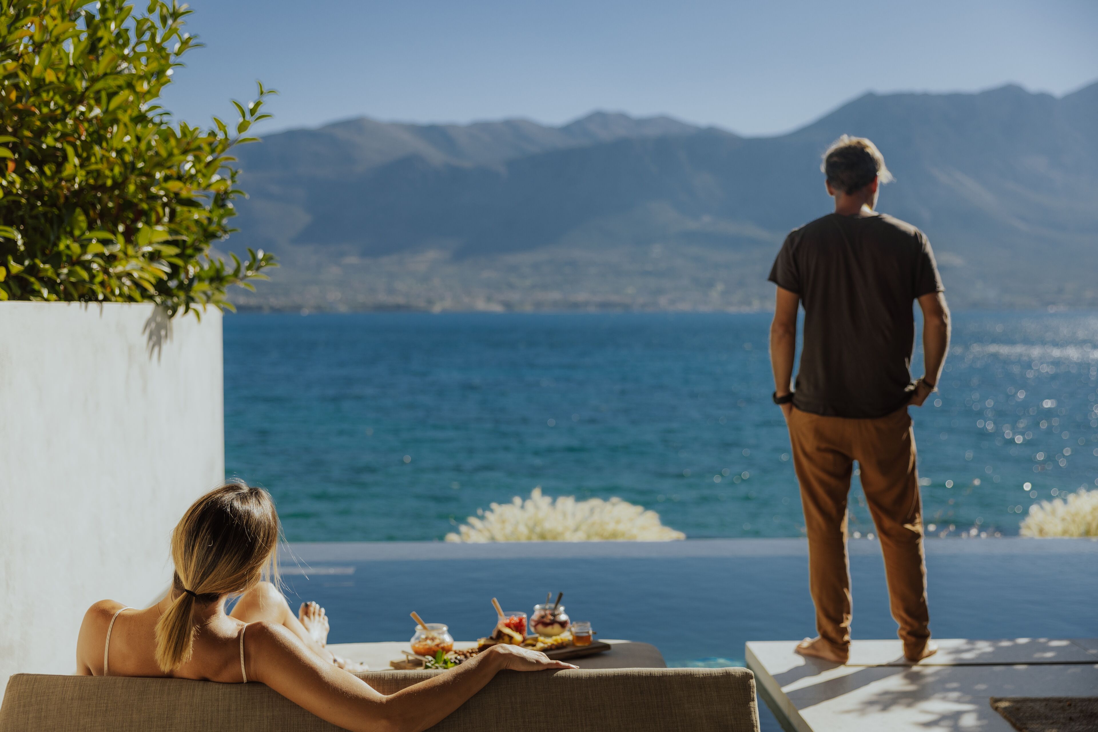 A couple relaxing by a pool with a view of the sea and mountains.