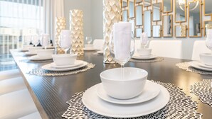 Een eettafel gedekt met witte borden, kommen en glazen, omringd door decoratieve vazen en een geometrisch wandspiegel.