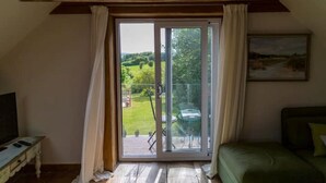 A room with a large sliding glass door leading to a balcony with a view of greenery. There is a bed with a green cover, a small table with a television, and a painting on the wall.
