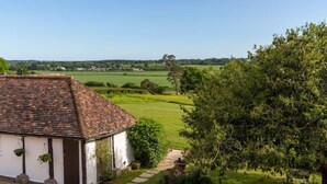 A white building with a tiled roof, a garden with a stone pathway, and a view of a green field and distant trees.