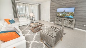 Una sala de estar moderna con un sofá blanco, un sillón gris y una mesa de centro. Un televisor de pantalla plana está montado en una pared gris, y hay una ventana con cortinas grises.