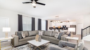 A modern living room with a sectional sofa, a coffee table, and a grey armchair. A kitchen is visible in the background with a dining area. - Last Minute Disney! Private Pool, Hot Tub, Game Room & Themed Bedrooms. Book Now! #49413 (Orlando)