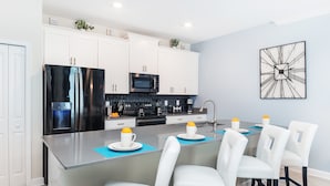 Une cuisine moderne avec des meubles blancs, un réfrigérateur noir, un four à micro-ondes et une machine à café. On y trouve également un coin repas avec des chaises blanches et un set de table bleu.