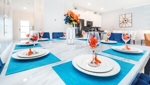 Un espace repas avec une longue table dressée pour accueillir plusieurs convives, agrémentée d’assiettes blanches, de sets de table bleus et de verres transparents décorés d’orange.