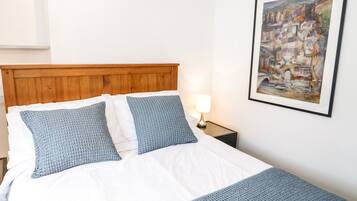 Un lit bien fait, avec une tête de lit en bois, deux oreillers, une couette blanche et un plaid bleu. Un tableau encadré est accroché au mur, et une petite table de chevet avec une lampe se trouve à côté du lit.