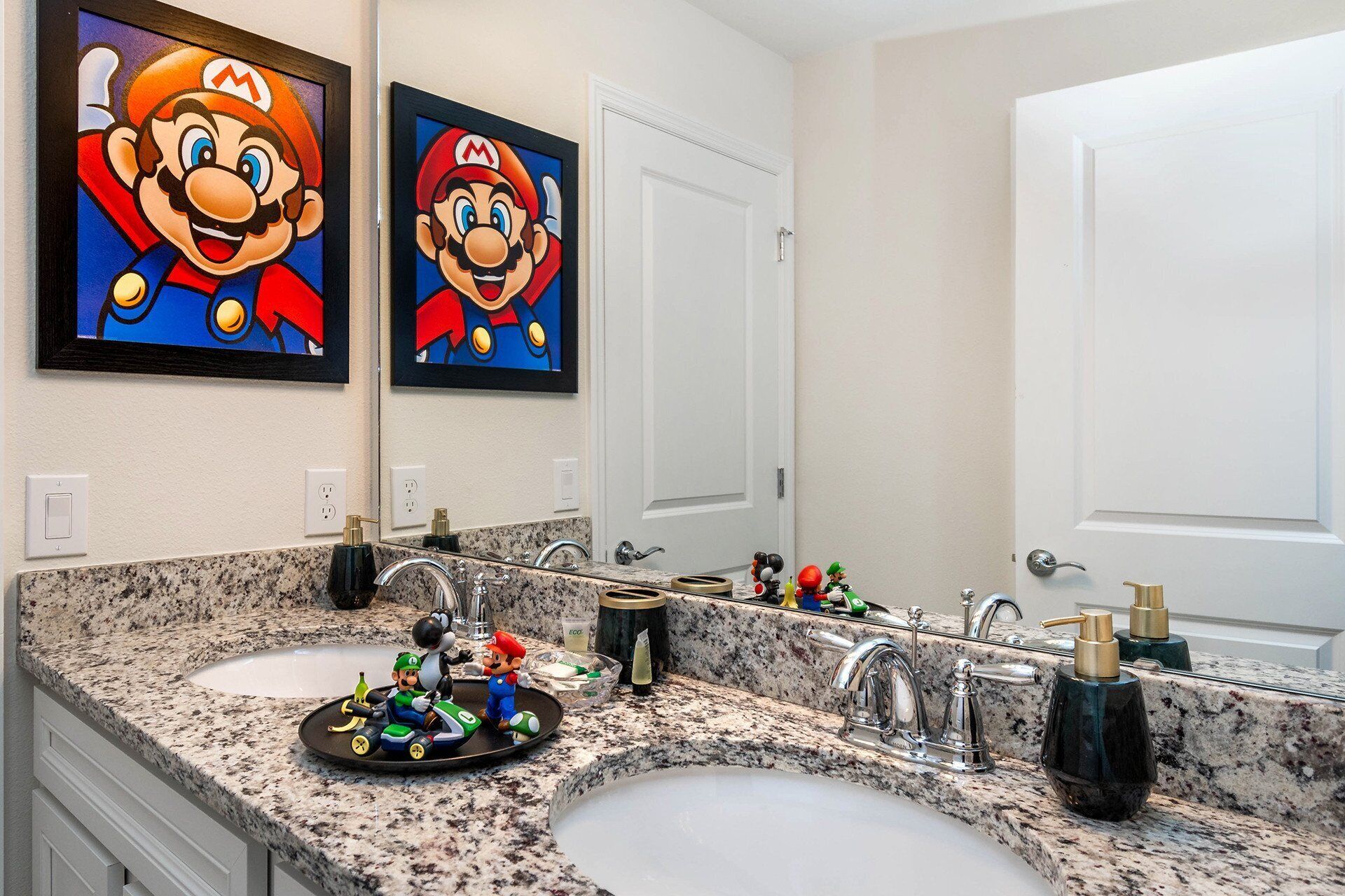 Une salle de bain avec deux lavabos, un miroir et des œuvres d’art encadrées représentant Mario.