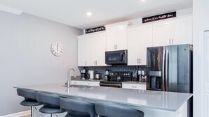 Una cocina moderna con armarios blancos, electrodomésticos de acero inoxidable, una isla central con taburetes y un reloj en la pared.
