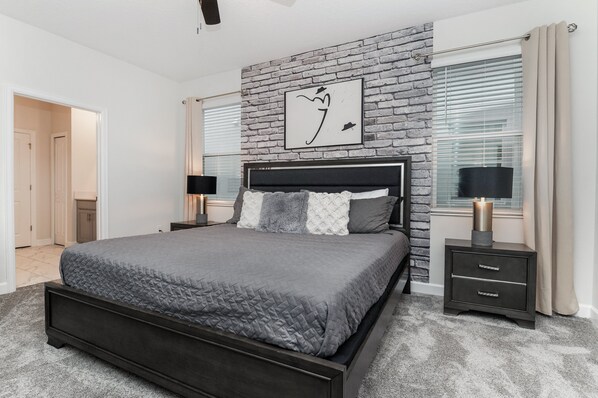 Un dormitorio moderno con una pared de ladrillo decorativa, una cama grande con cubrecama gris acolchado, dos mesitas de noche con lámparas y una obra enmarcada sobre la cama.