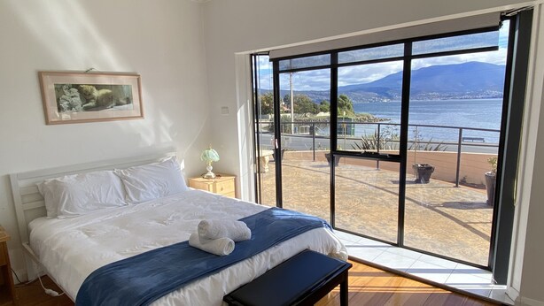 Un dormitorio con un ventanal grande, una cama con ropa de cama blanca, un plaid azul y un banco.