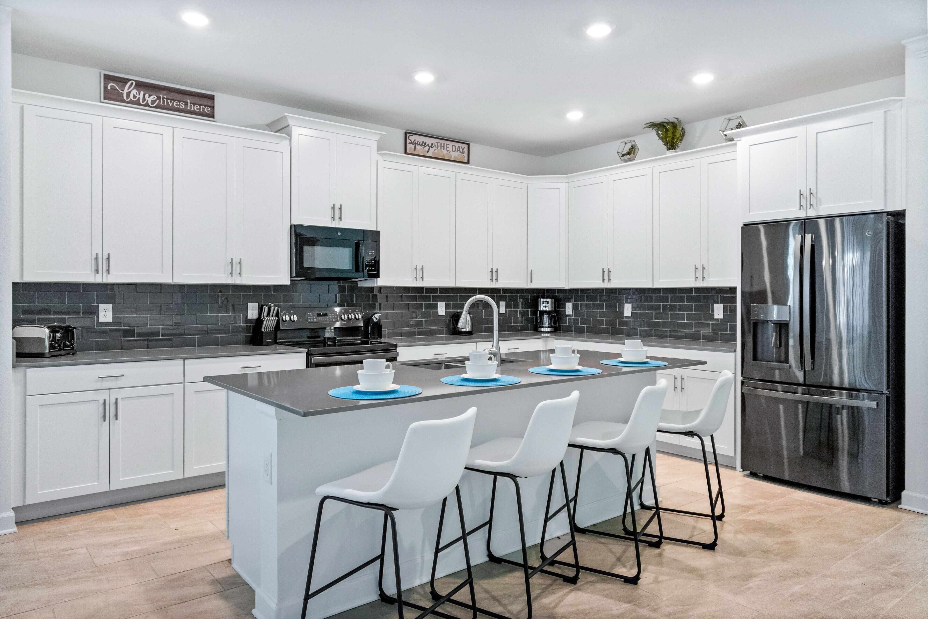 A modern kitchen with white cabinets, a stainless steel refrigerator, a central island with bar stools, and a backsplash of dark tiles.