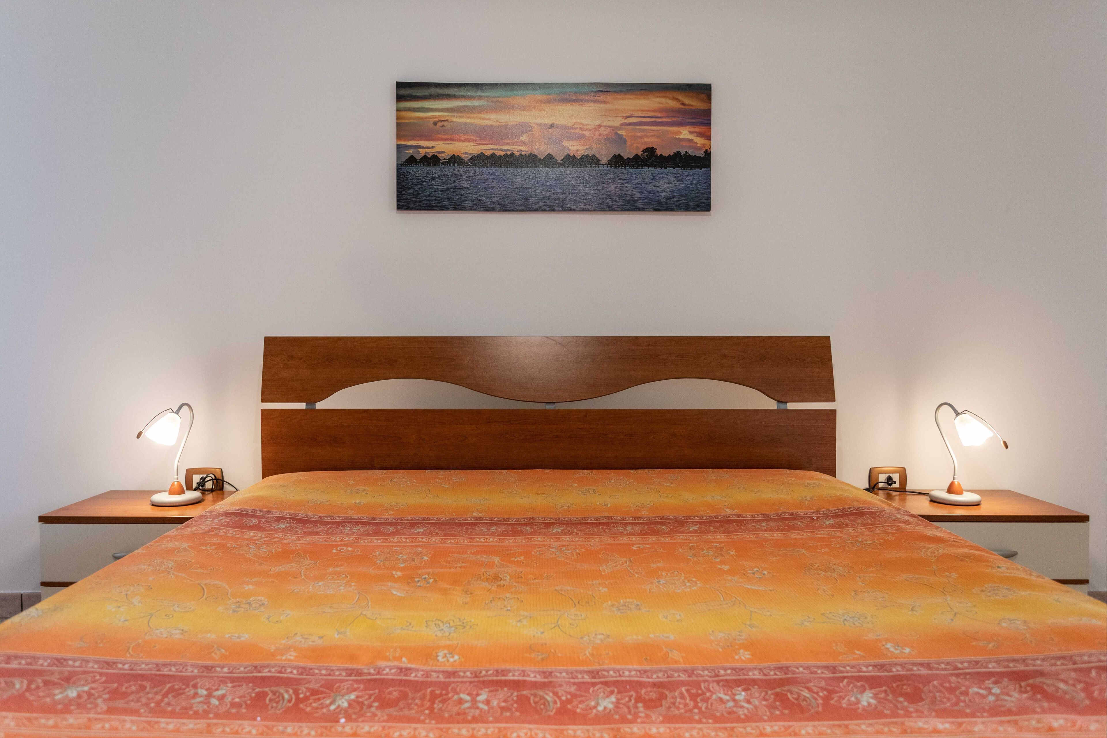 A bedroom with a bed, two bedside tables, a nightstand, a wooden headboard, and a painting of a sunset over water on the wall.