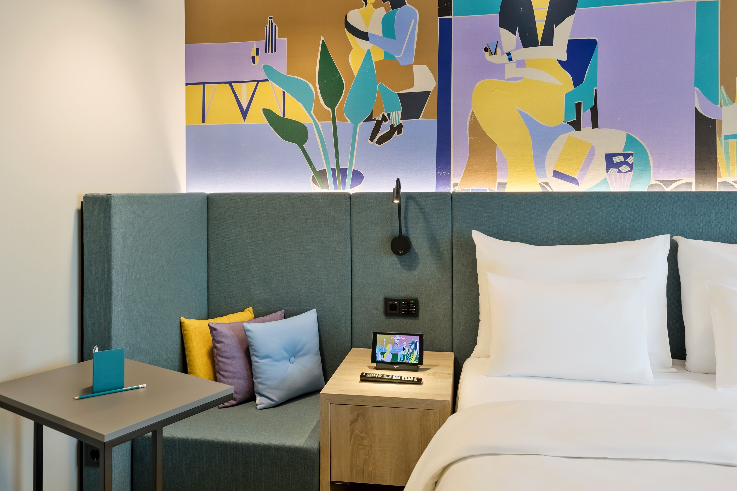 A modern hotel room with a bed, a bedside table with a laptop, a small desk with a chair, and a colorful abstract painting on the wall.