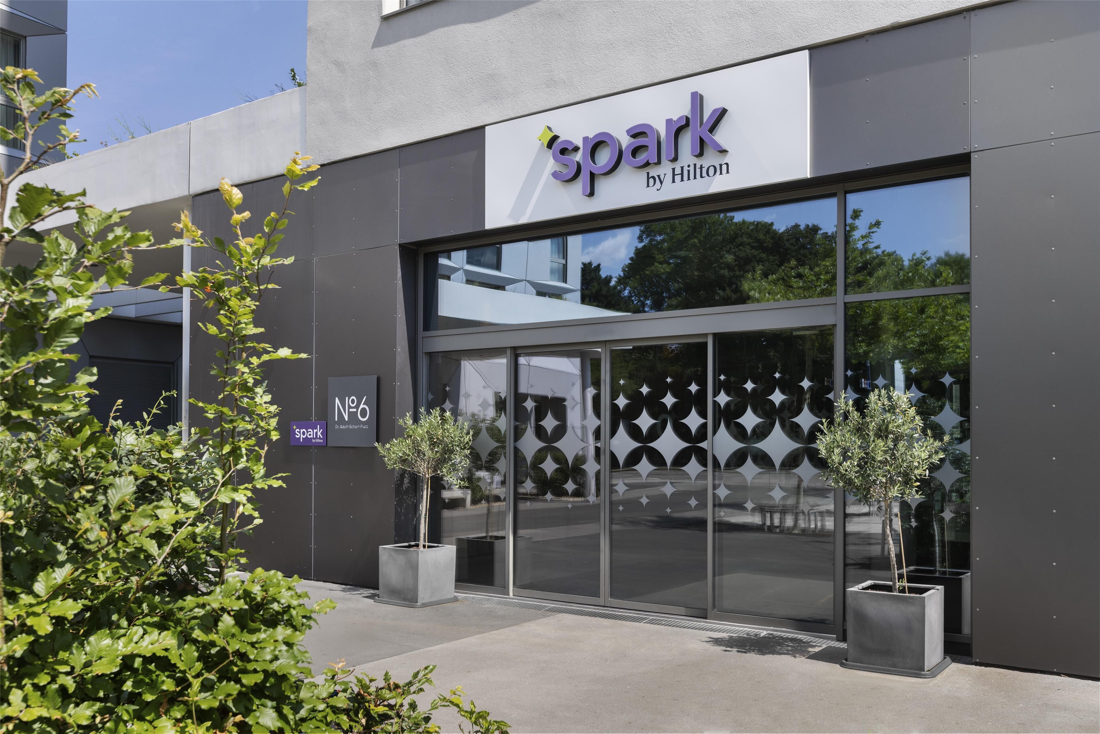 A hotel named 'Spark by Hilton' with a modern facade, large glass windows, and two potted plants in front.