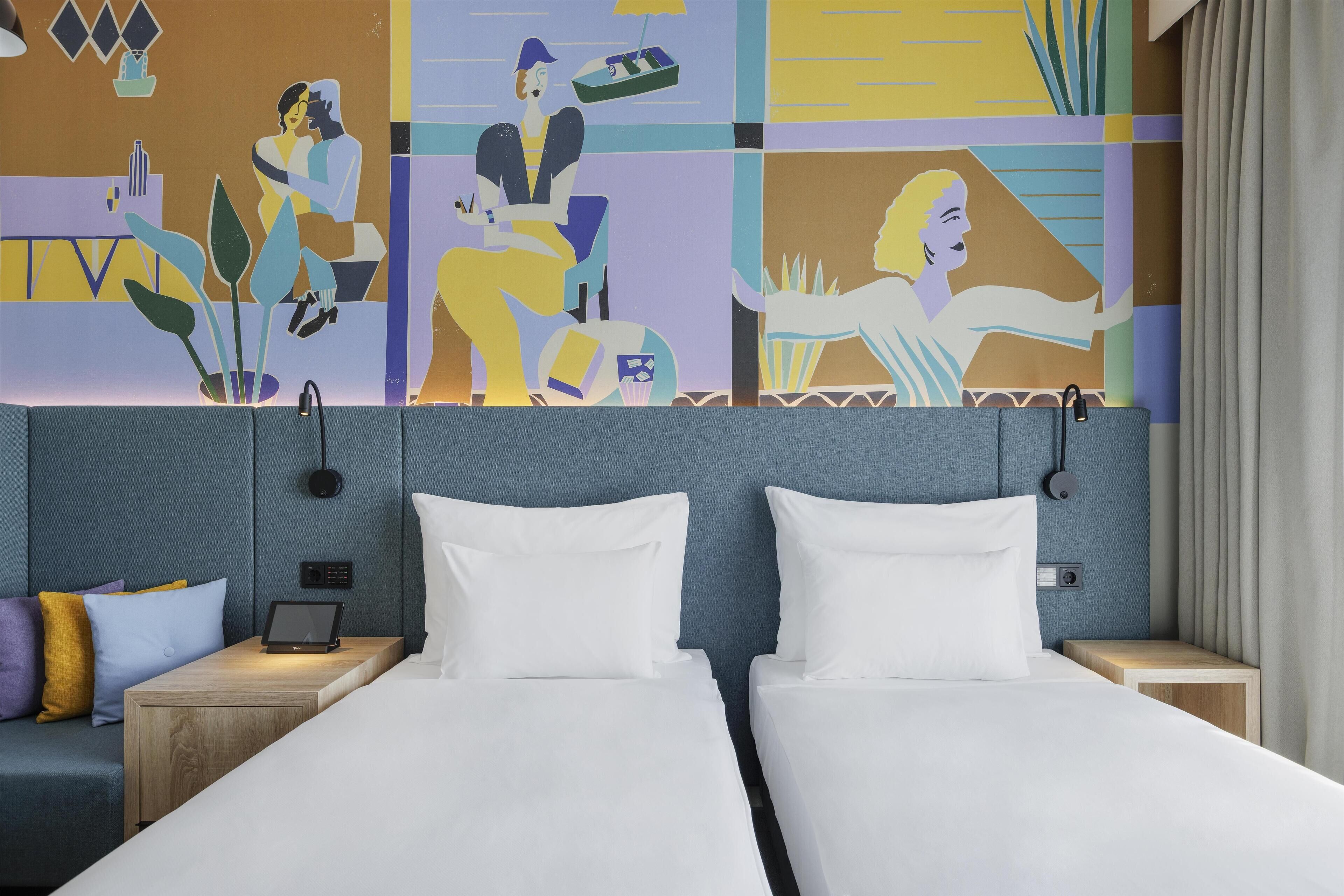 A hotel room with two beds, a desk, a lamp, and a colorful abstract mural on the wall.