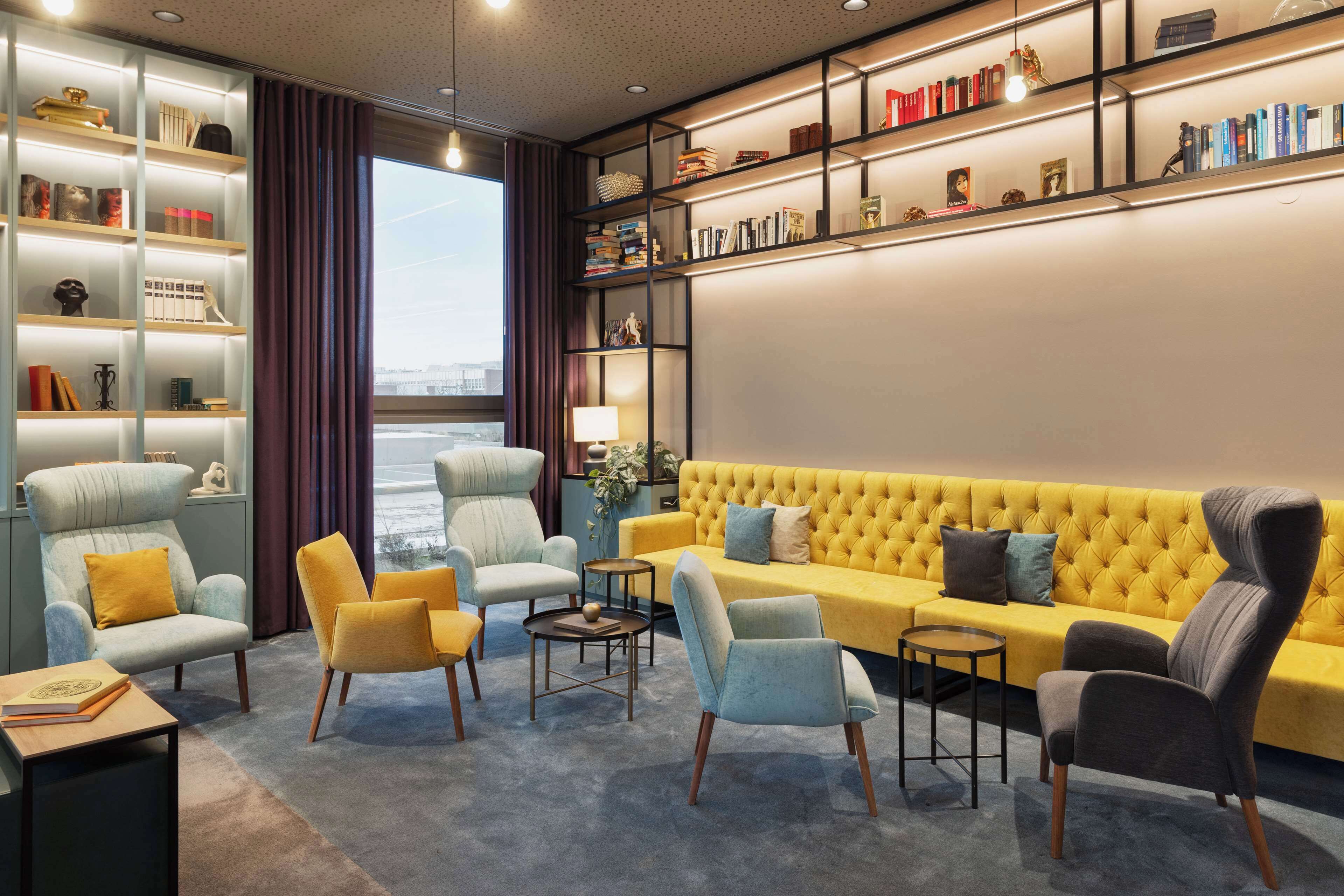 Una sala de estar moderna con un sofá amarillo, sillones azul claro y amarillos, una estantería y vistas a través de un ventanal.
