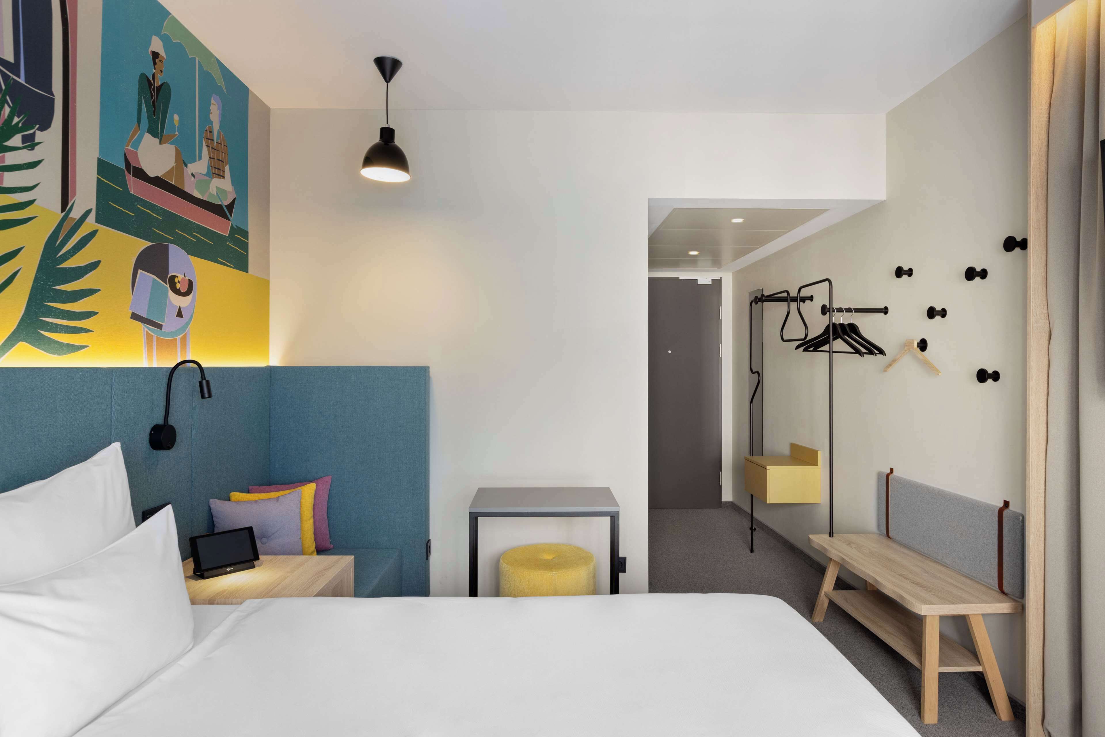 A modern hotel room with a bed, bedside table, and a small seating area. There is a wall-mounted mirror and a door leading to another room.