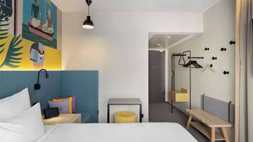A modern hotel room with a bed, bedside table, and a small seating area. There is a wall-mounted mirror and a door leading to another room.