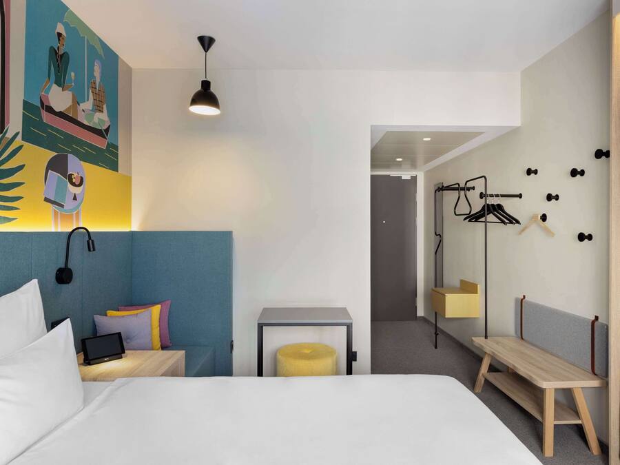 A modern hotel room with a bed, bedside table, and a small seating area. There is a wall-mounted mirror and a door leading to another room.