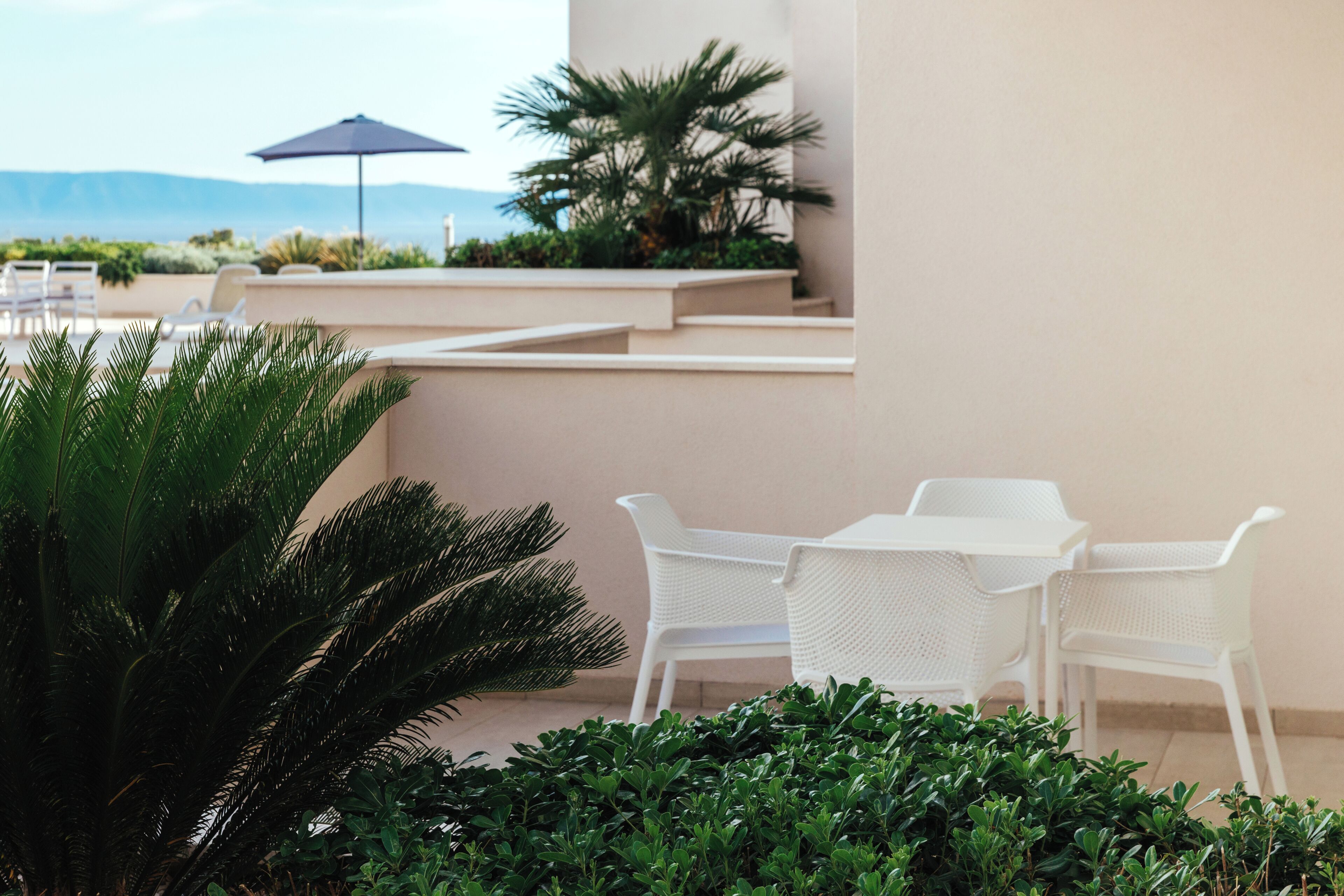 Una terraza con muebles de mimbre blanco, una mesa y sillas, rodeada de vegetación y vistas a las montañas.