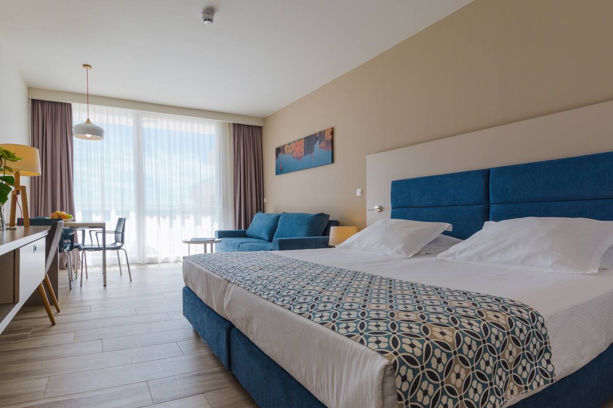 Een hotelkamer met een groot bed, een bureau met stoel, een bank en een groot raam met gordijnen.
