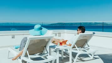 Två personer som kopplar av i vita solstolar vid en pool, med fri utsikt över havet och de avlägsna kullarna.