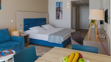 A modern hotel room with a bed, a dining table with fruit, a sofa, and a lamp.
