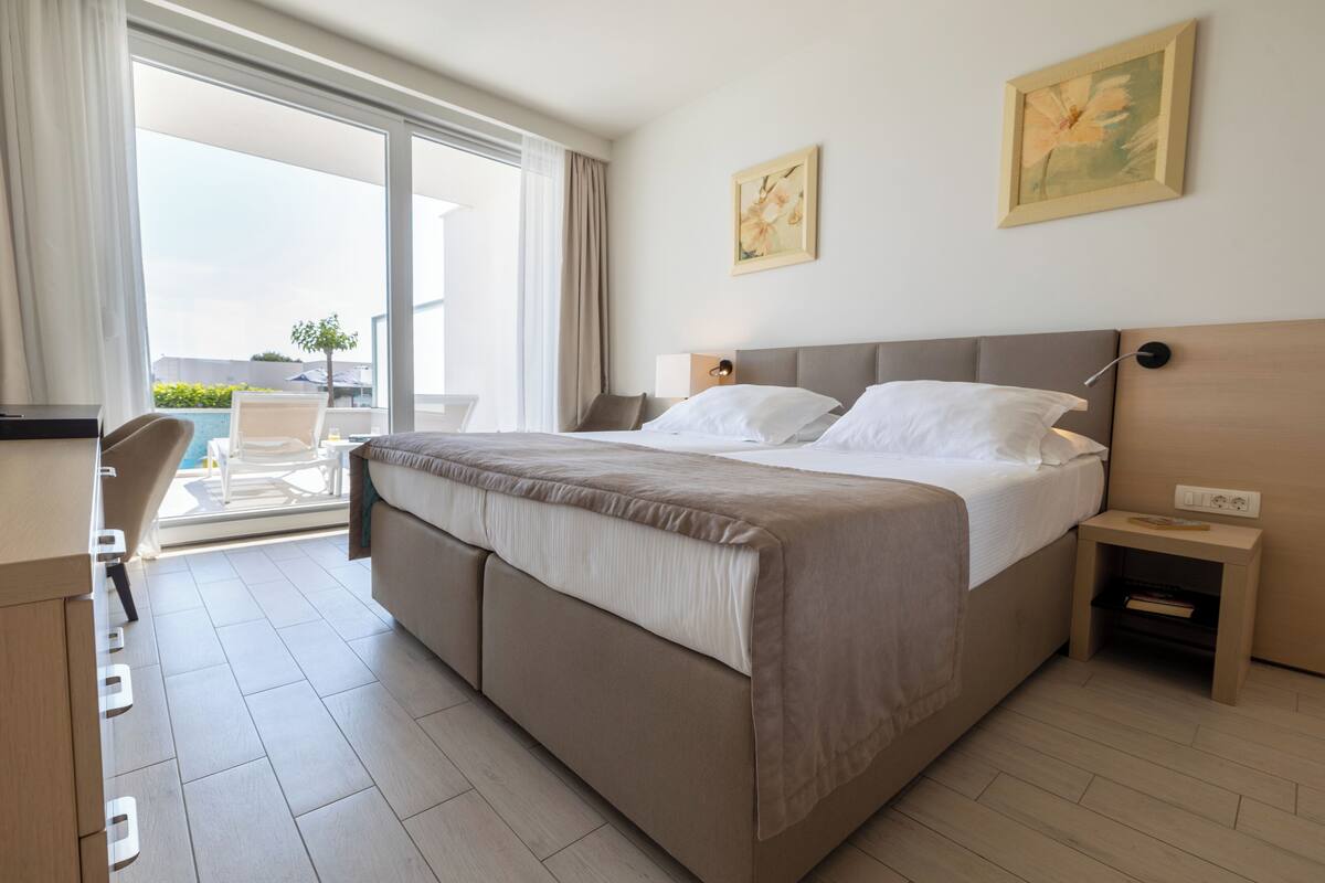 Een hotelkamer met een groot bed, een bureau en een stoel. Er is een raam met gordijnen, een schilderij aan de muur en een klein tafeltje met een lamp.
