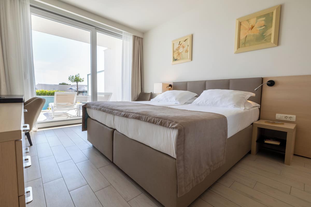 Une chambre d’hôtel avec un grand lit, un bureau et une chaise. Il y a une fenêtre avec des rideaux, un tableau au mur et une petite table avec une lampe.