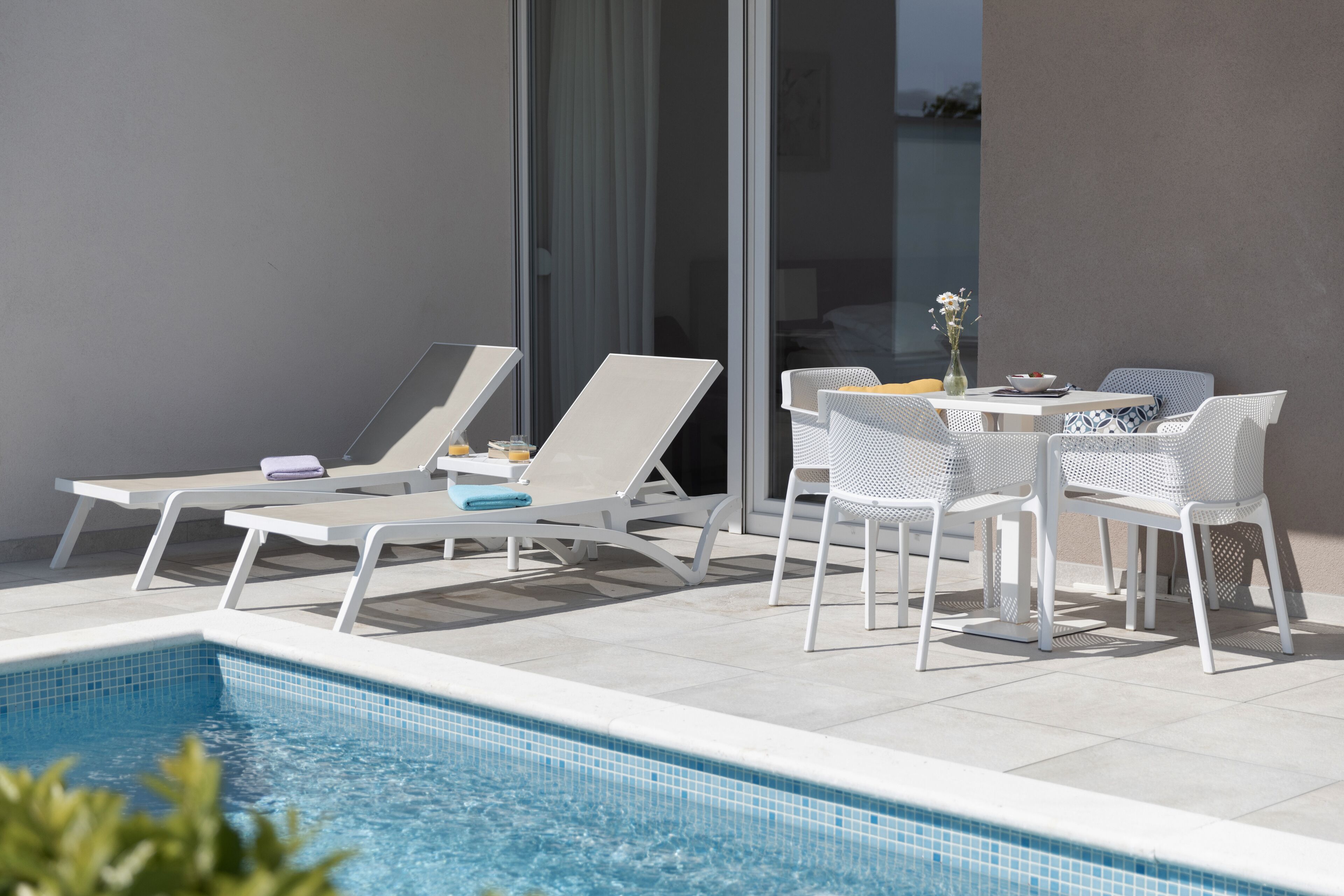 Area piscina con lettini e un tavolo, una piscina blu cristallina e vista sul mare attraverso ampie finestre.